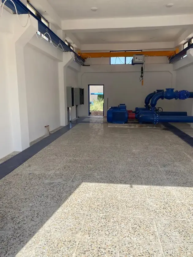 Interior view of a water pumping station with blue pipes, pumps, and electrical panels.