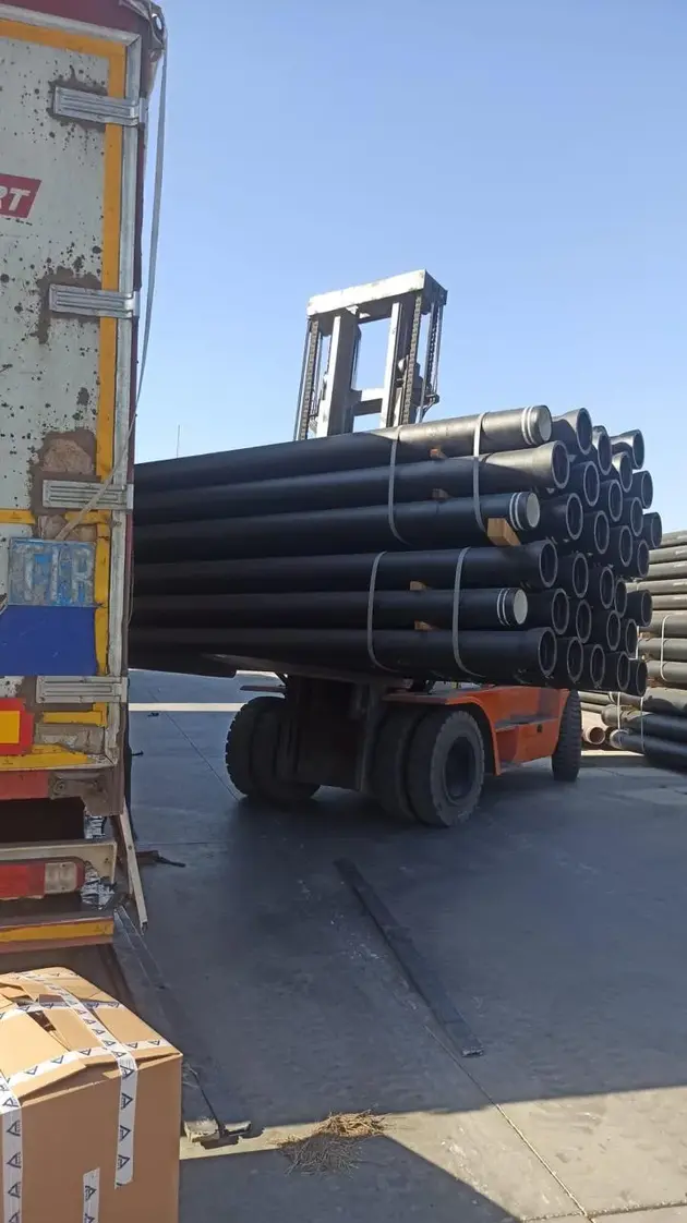 A forklift carries a large bundle of black pipes, loading them onto a truck at a construction materials yard.