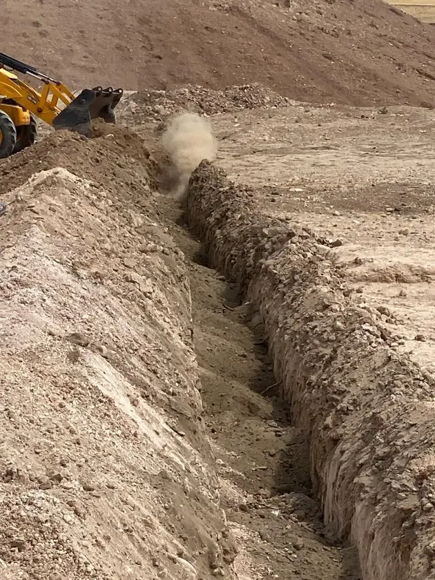 A yellow excavator digging a long trench in a dry, dusty landscape, with a cloud of dust rising from the excavation.